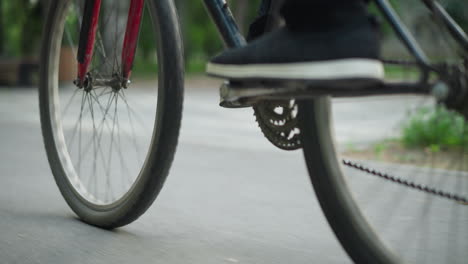 rear view of someone wearing sneakers standing while riding a bicycle on a paved road, captured from a low angle, the background shows blurred elements of greenery and objects