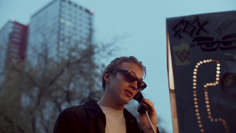 young stylish man having conversation at phone booth in evening
