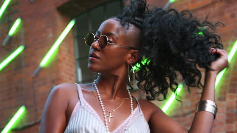 joven negra de moda con gafas de sol redondas jugando con su cabello, sonriendo y levantando gafas de sol, ángulo bajo, primer plano, luces borrosas de neón en el fondo