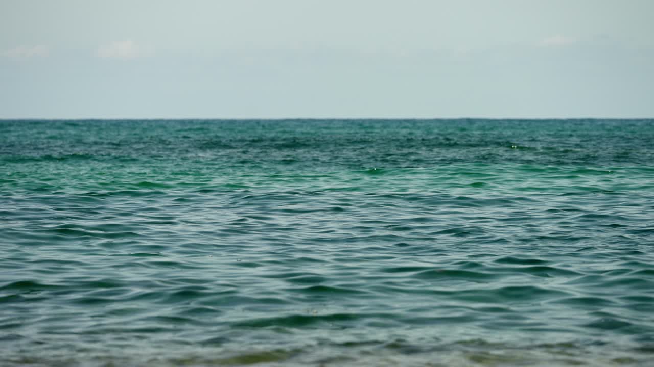 Calm turquoise ocean waves move gently in extreme slow motion, filmed from a low beach angle with a peaceful tropical atmosphere.