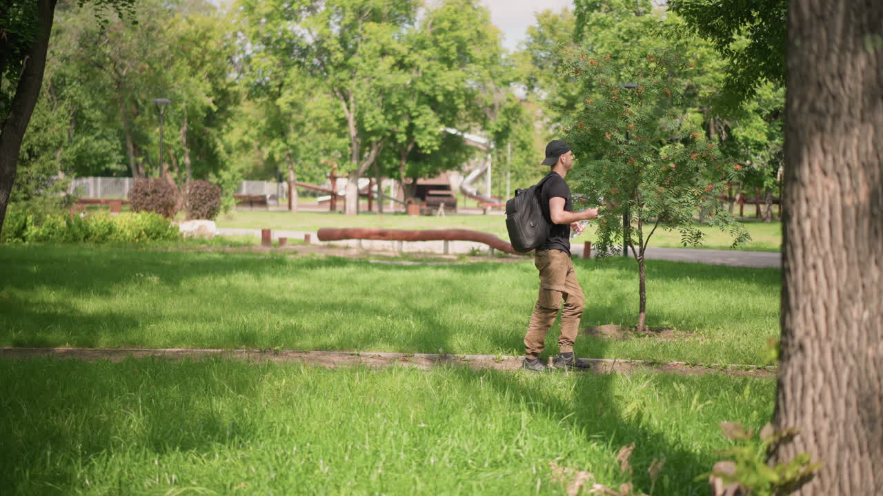 White Male Student Walking Through Park Backpacked Along Leafy Path, Casual Stride Past Trees And Benches, Summer Sunlight, Smartphone Glance, Relaxed Campus Mood, Green Grass And Distant Pond