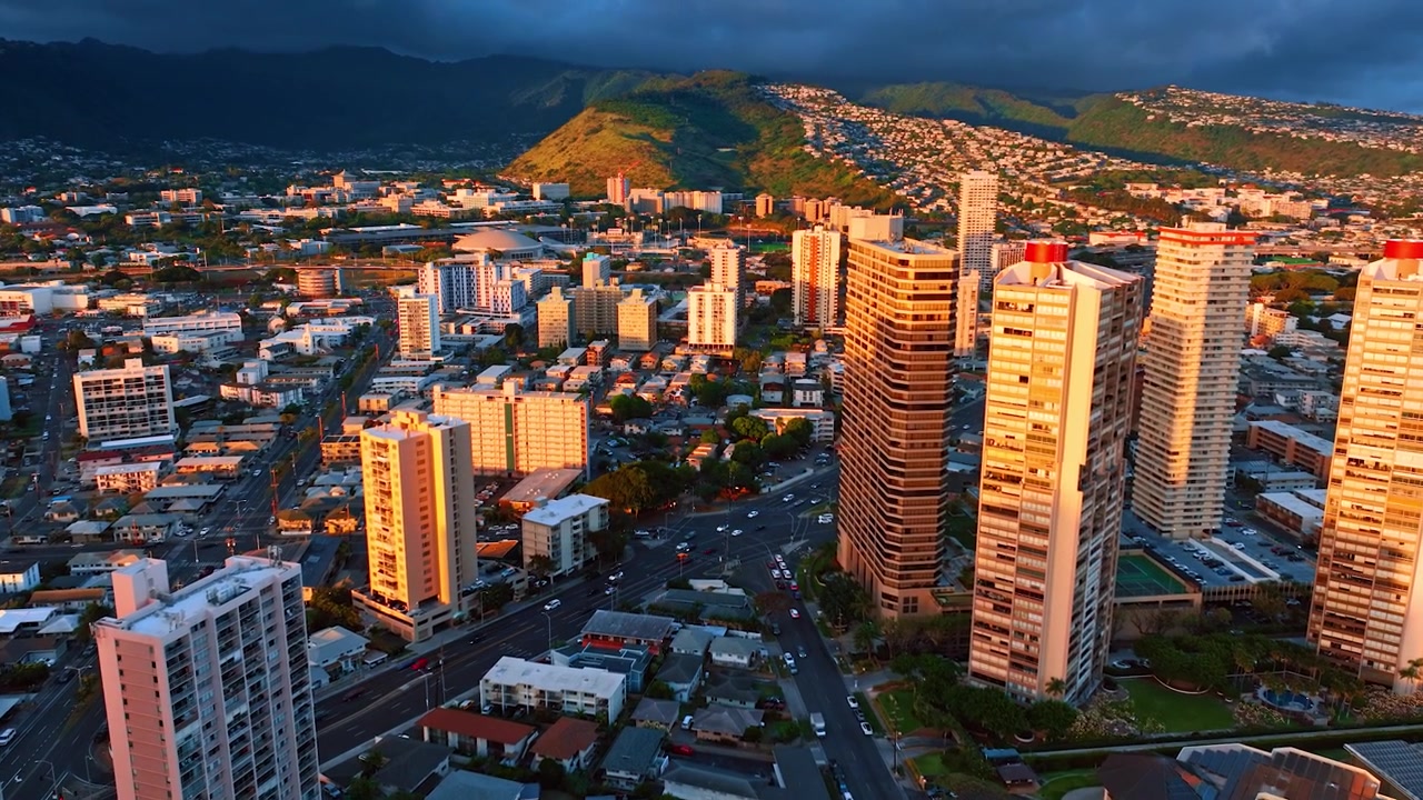 Grattacieli di Honolulu nella luce arancione del tramonto. Diverse auto si muovono lungo la strada che attraversa la città. Montagne verdeggianti sono coperte da nuvole scure sullo sfondo