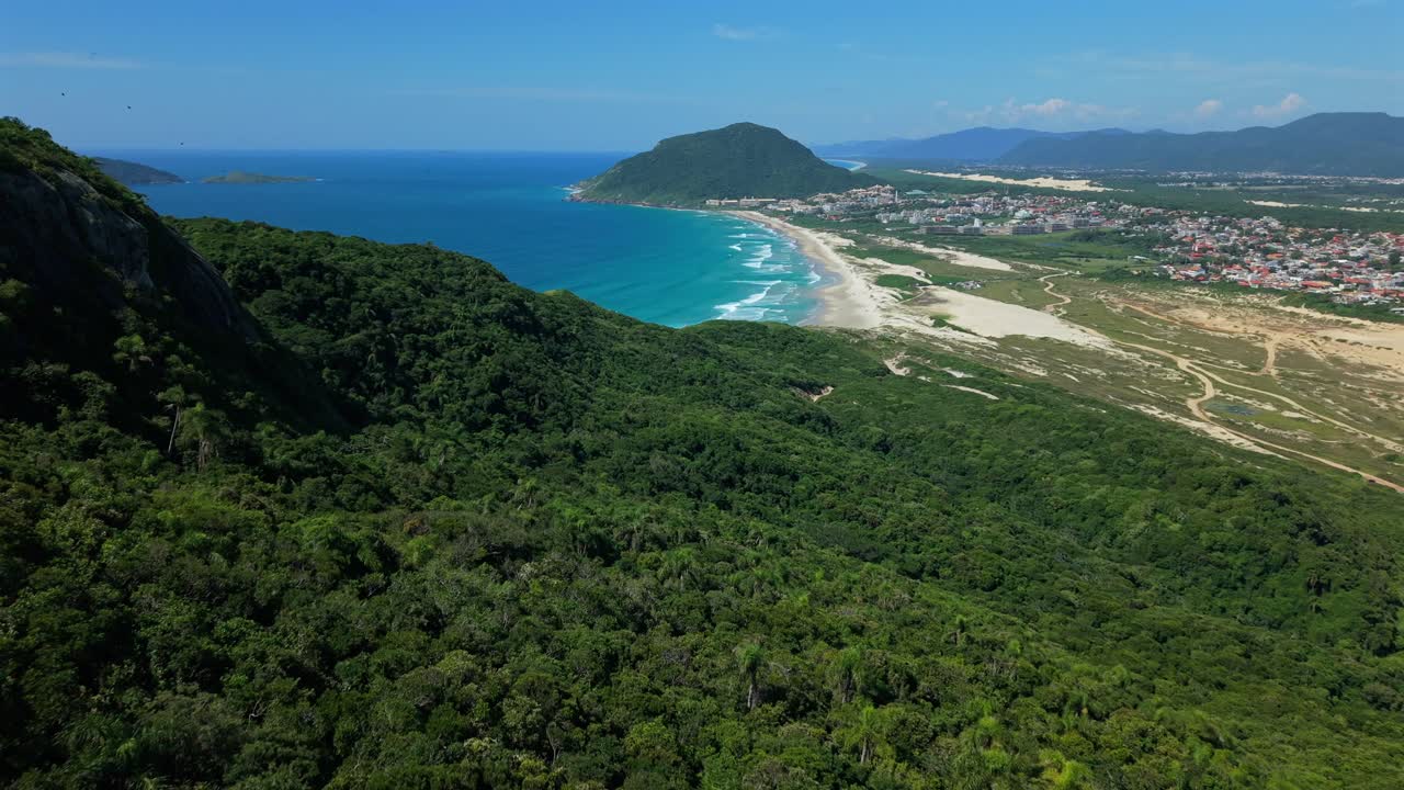 Drone che sorvola una lussureggiante foresta montana rivelando una vista panoramica della costa della spiaggia di Santinho, delle dune di sabbia e di una città tropicale a Santa Catarina, Brasile