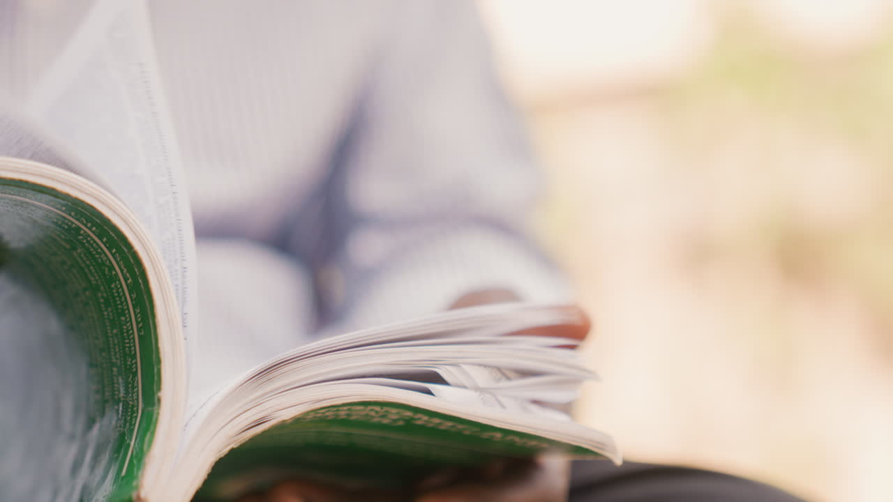 Mani che sfogliano pagine di rivista alla luce del sole. Sfondo bokeh soffuso, manica di camicia casual, primo piano ravvicinato su mani e pagine verdi lucide aperte, giri lenti e intenzionali, atmosfera rilassata del fine settimana, caffè all'aperto