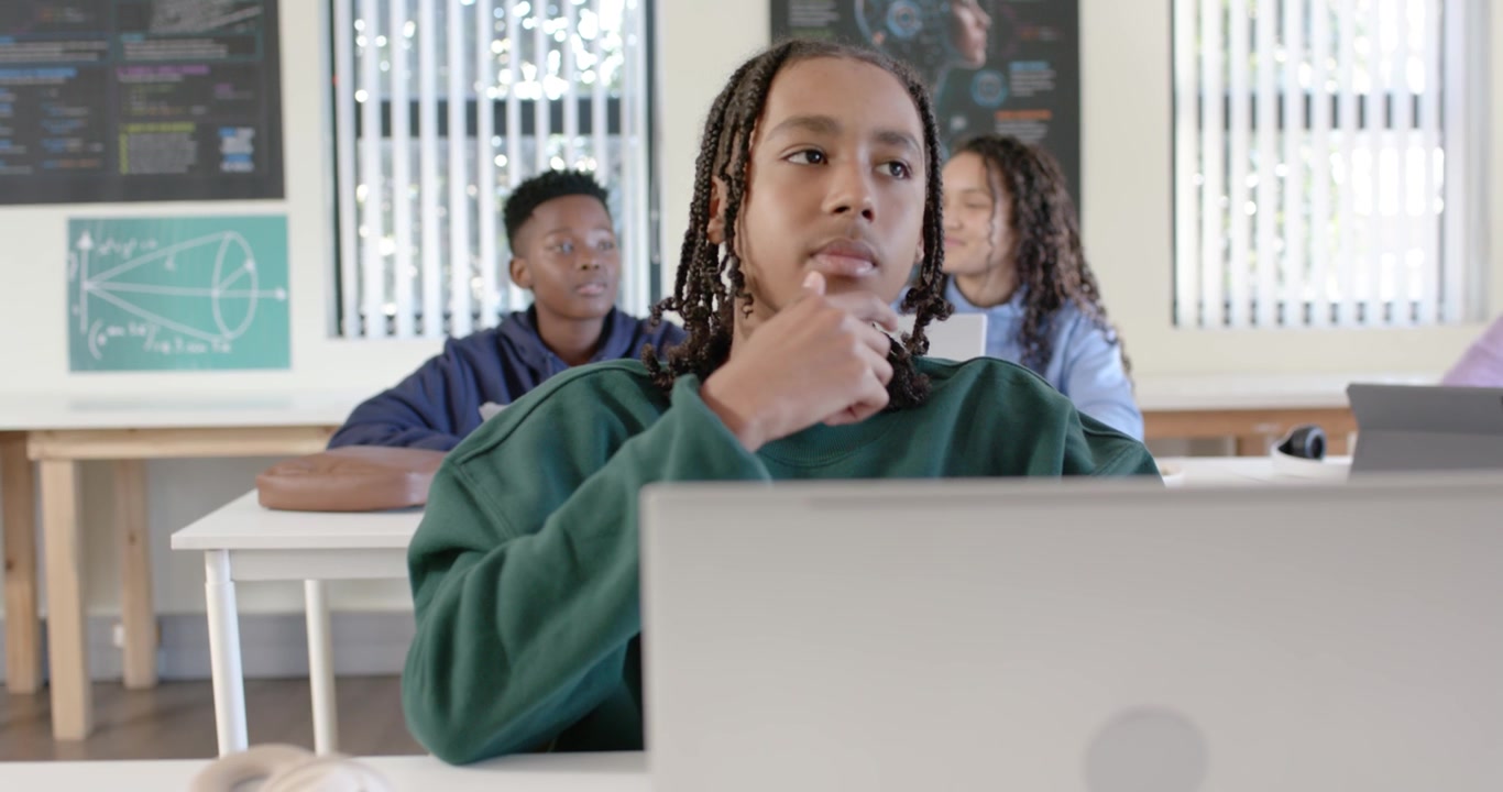 Teacher walking class aisles causing teen male shifting gaze toward laptop and peers, resting chin