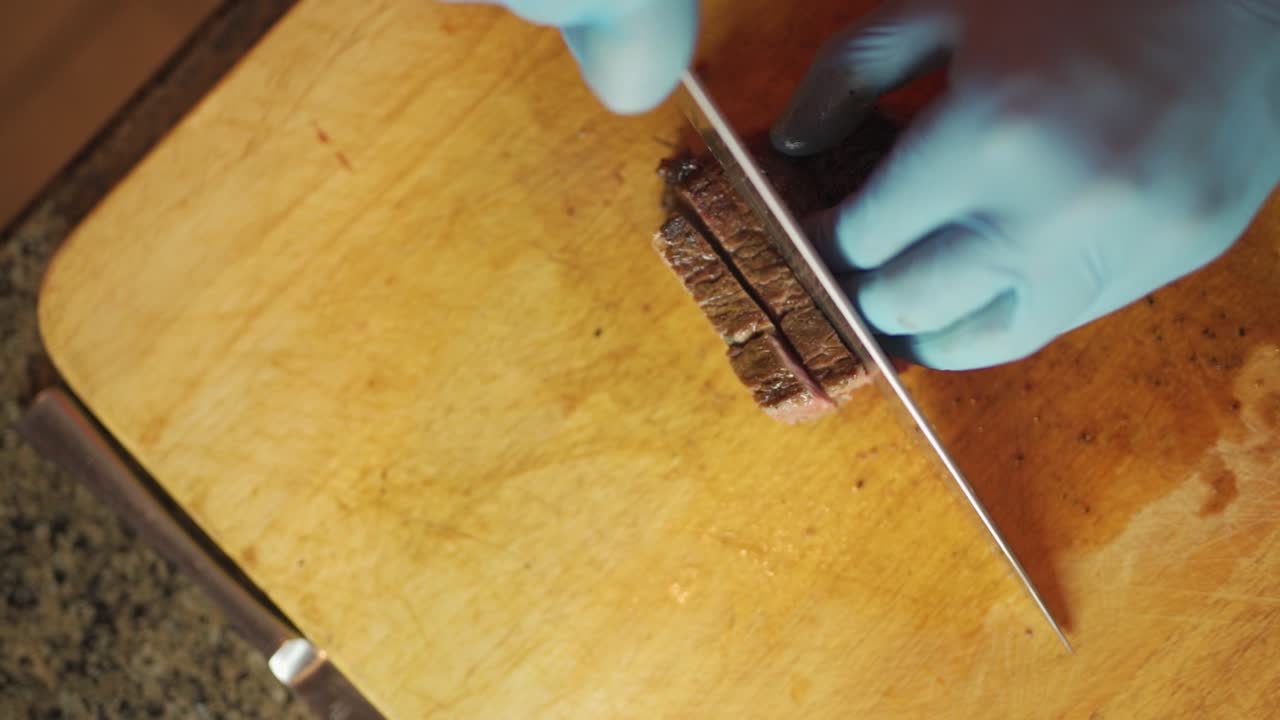 Circling footage of a food preparer, observing sanitary procedures, using a large carving knife to make cuts out of a juicy roast on a wooden cutting board.