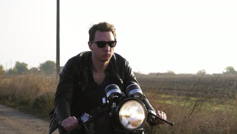 closeup view of a stylish cool young man in sunglasses and leather jacket riding motorcycle on a asphalt road on a sunny day