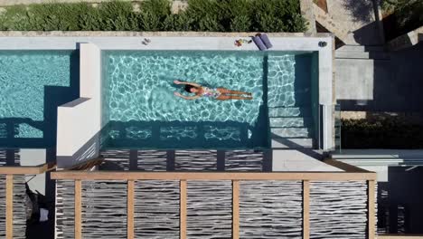 Brunette-woman-in-swimming-costume-enjoying-the-pool-at-CayoResort-in-Greece-Crete---she-swims-on-her-back-and-enjoys-the-water---drone-flies-in-bird's-eye-view