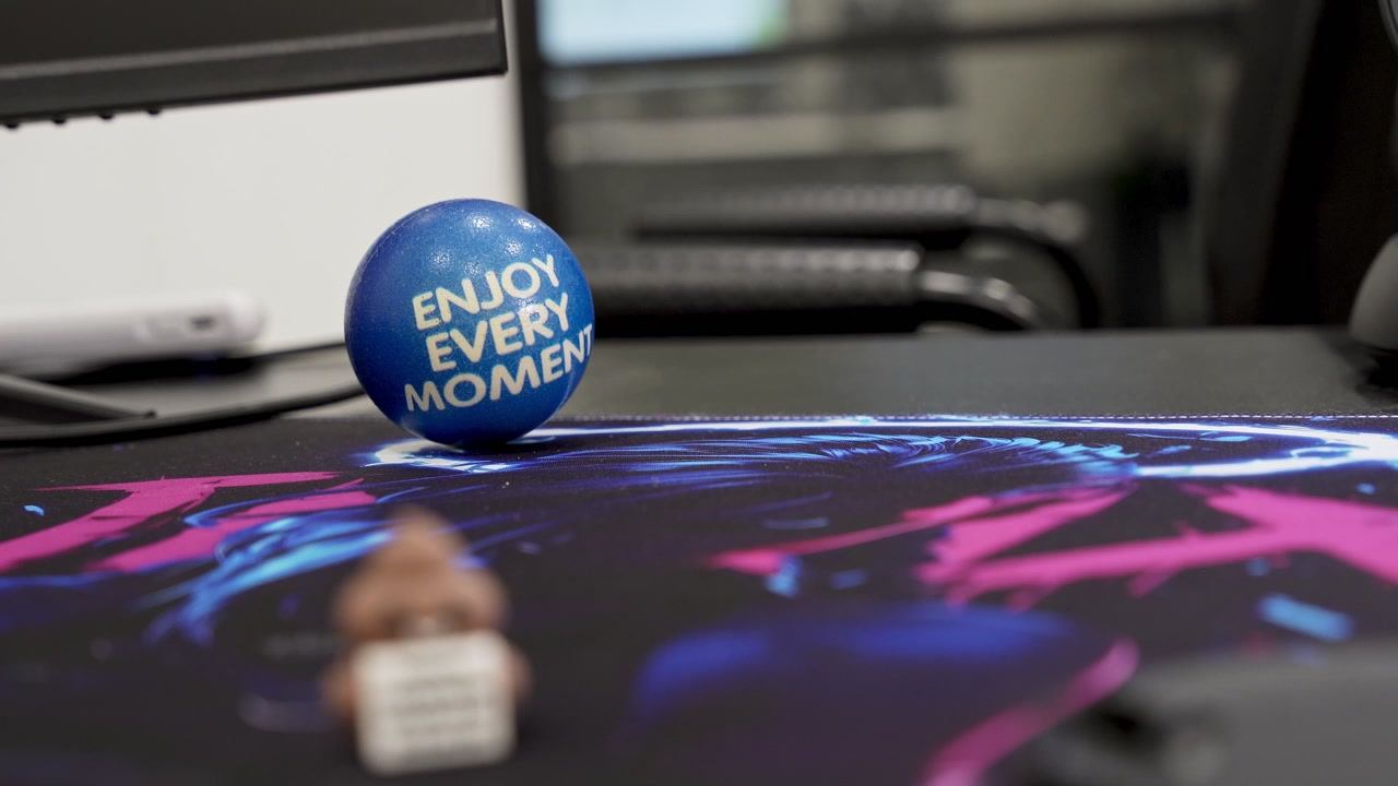 Close-up of a blue motivational stress ball reading Enjoy Every Moment on a colorful desk mat in a modern computer workspace.