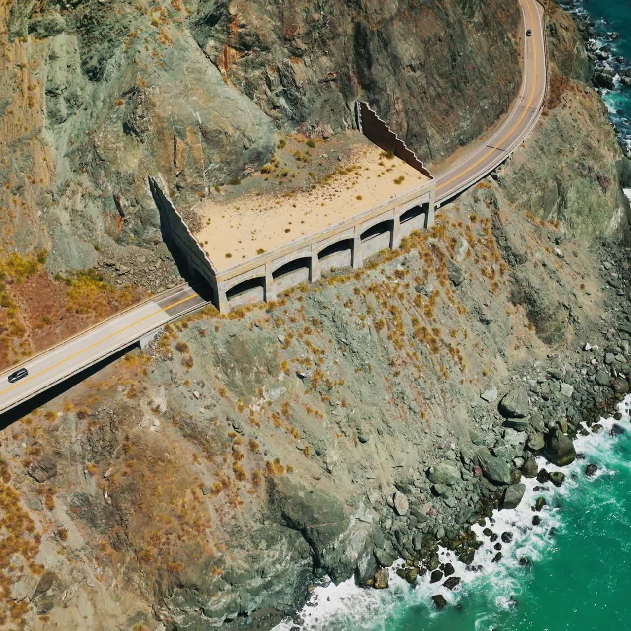 Cars moving by the dangerous road in the mountains of pacific shore. Turquoise ocean waves crush by the rocks. Top view