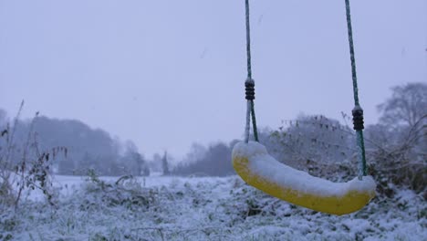 Eine-Schöne,-Ruhige,-Kühle-Zeitlupenaufnahme-Einer-Schaukel,-Die-Sich-Bei-Schnee-Sanft-Im-Wind-Im-Vordergrund-Wiegt,-Mit-Einem-Weißen,-Schneebedeckten-Feld-Im-Hintergrund