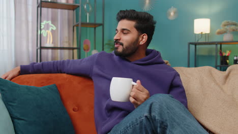 handsome smiling indian man drinking a cup of coffee or herbal tea sitting at home sofa, break time