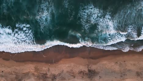 overhead view of the waves on a beach with orange sand at sunset with people walking on the shore