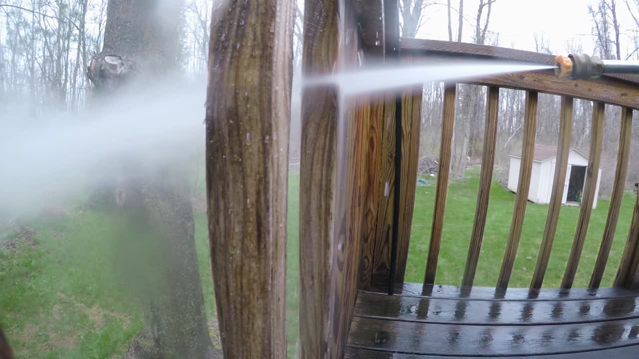 A man uses a power washer to clean a wooden deck railing post