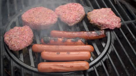 el primer plano de la parrilla al aire libre mientras las hamburguesas de ternera sazonadas y los perritos calientes se vuelven llamas con humo