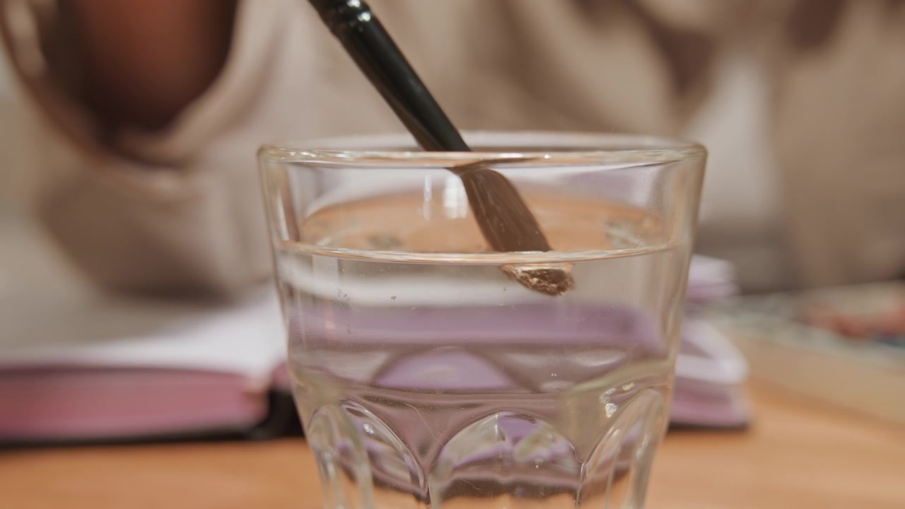 Primo piano di un bicchiere pieno d'acqua posizionato su una superficie di legno. Una persona intinge e mescola un pennello nel liquido, creando increspature. Un libro aperto è visibile sullo sfondo. L'illuminazione soffusa evidenzia la texture chiara