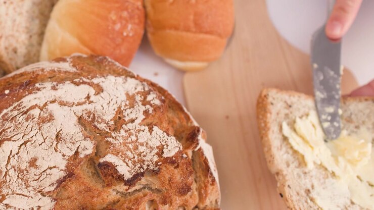 Close-up overhead shot of a hand spreading butter on multigrain bread beside sourdough loaves.