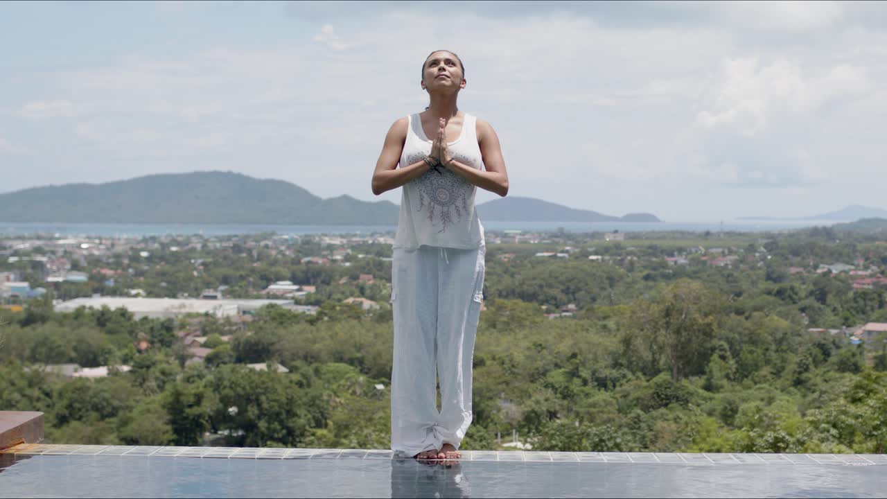 Mulher meditando em pé à beira da piscina com a costa tropical ao fundo