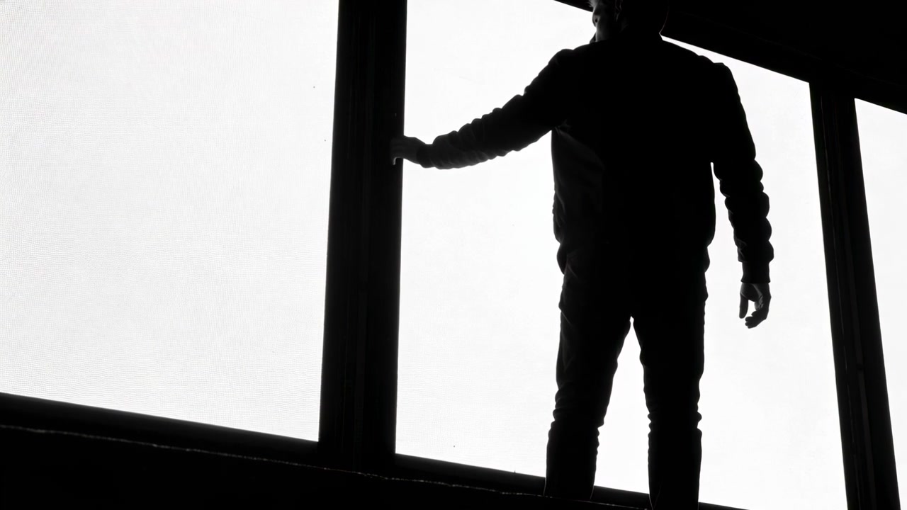 Walking backlit man entering left slowing in hall touching mullion at window in jacket gazing out