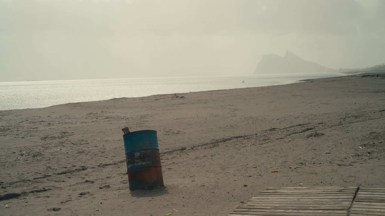 Barile di metallo arrugginito su una spiaggia sabbiosa con la Rocca di Gibilterra in lontananza sotto una luce caliginosa e drammatica.
