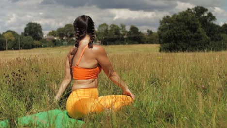 woman sitting in long grass field doing yoga, stretching arms up to side bend to easy pose bound hands