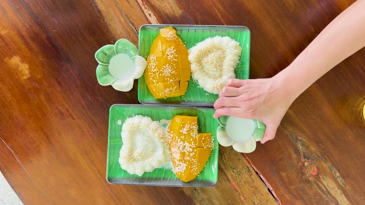 A hand pours coconut milk over mango sticky rice on a wooden table in natural light, creating a vibrant and appetizing scene
