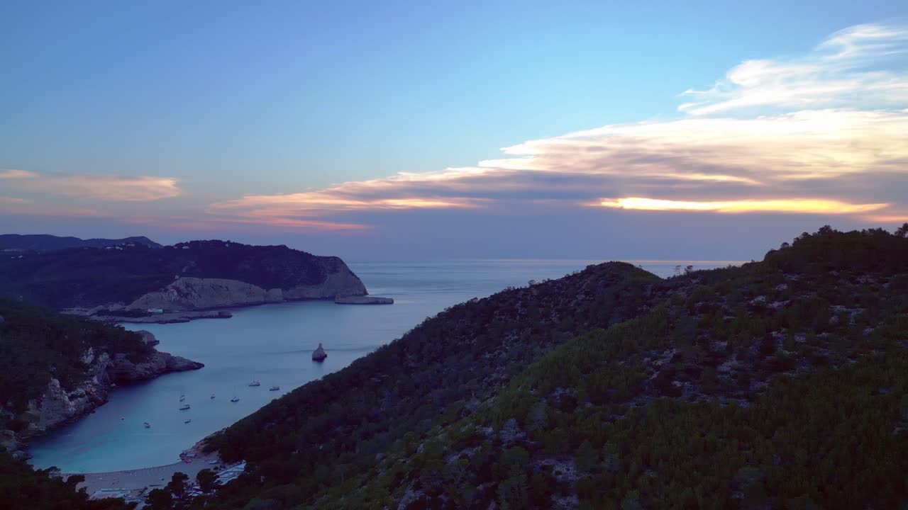 Panoramisk udsigt over Cala Benirras-bugten og skovklædte bakker i skumringen på Ibiza. Drone roterer og panorerer til venstre