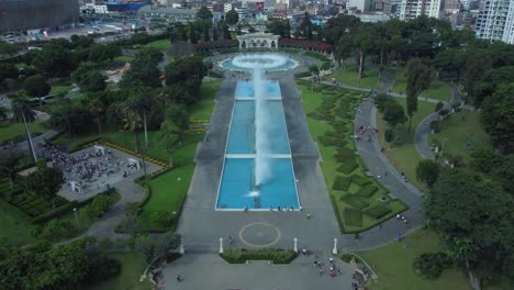 Drone-flies-over-a-rectangular-water-fountain,-towards-the-end-camera-tilts-down-on-a-circular-water-fountain