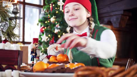 tricky female elf sitting at table on christmas tree background. girl in elf suit is taking delicious food on table