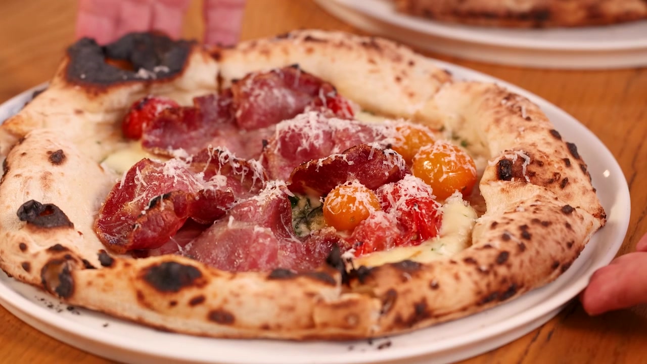 Uno chef ruota una pizza gourmet a lievitazione naturale guarnita con prosciutto e parmigiano su un tavolo.