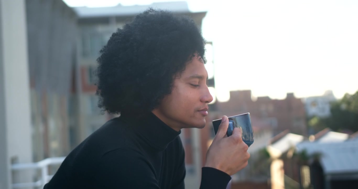 Afroamerikansk mand sidder på altanen i rullekrave, betragter byen og nipper til en blå keramisk kop.