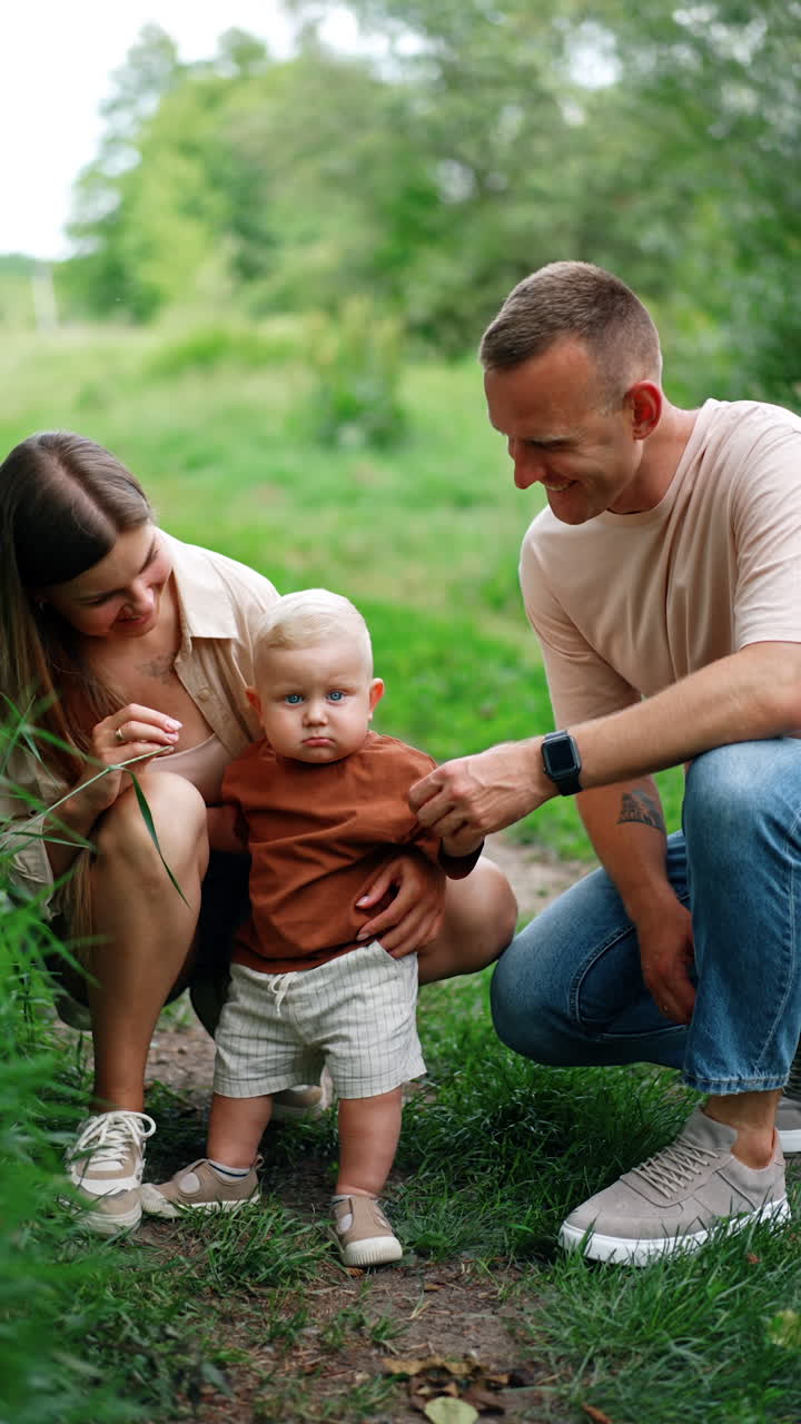 Loving Caucasian parents stand near their little child. Little toddler baby stands looking at camera attentively. Green nature at backdrop Vertical video.
