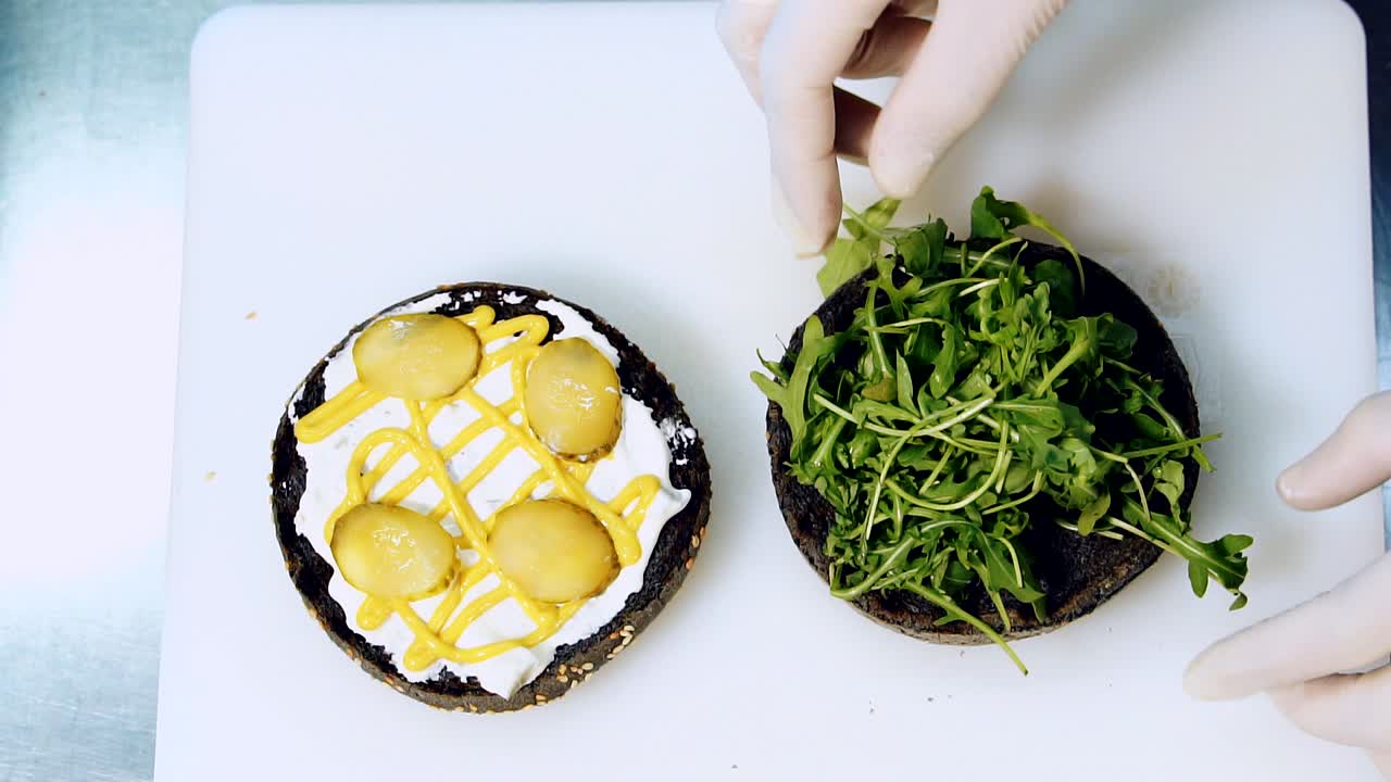 Cooking vegetarian burger. Hands of a chef putting green salad leaves on black bun. Culinary preparation of vegan hamburger. Close-up
