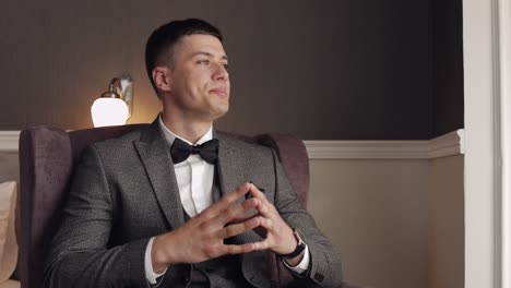 a man in a suit and bow tie sits in a chair and looks away.