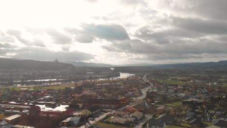 Rechts-Rotierende-Luftdrohne-über-Dem-Rand-Einer-Kleinen-Stadt-Mit-Silhouette-Einer-Burg-Auf-Einem-Hügel-Im-Hintergrund-Mit-Hohen-Wolken-Und-Fluss