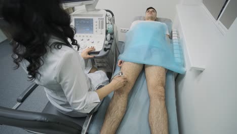 woman doctor hands examines young man patient knee-joint using ultrasound sensor device, sonography. doctor runs ultrasound sensor over patient's leg, knee.