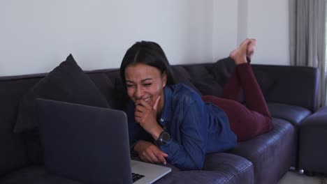 Mixed-race-woman-lying-on-couch-using-laptop-and-laughing