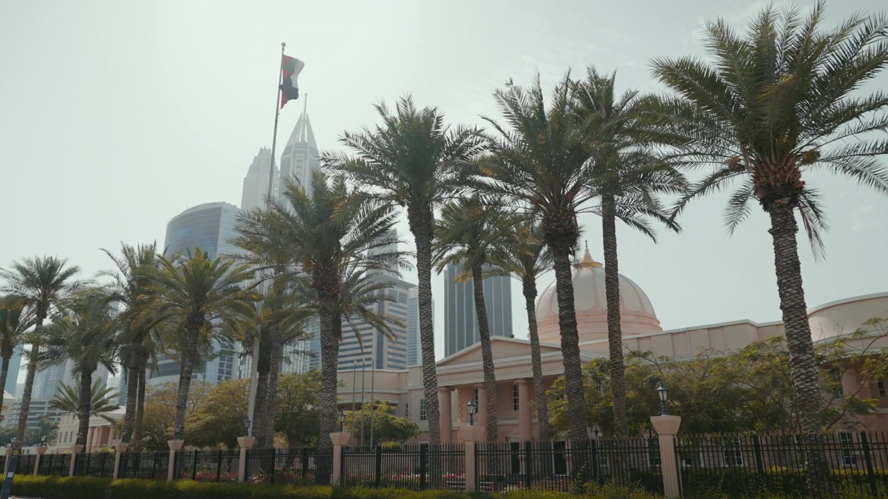 Ampia veduta dell'Università Americana di Dubai, che mostra il suo edificio, le palme circostanti, la bandiera degli Emirati Arabi Uniti e l'area di Media City e Marina.