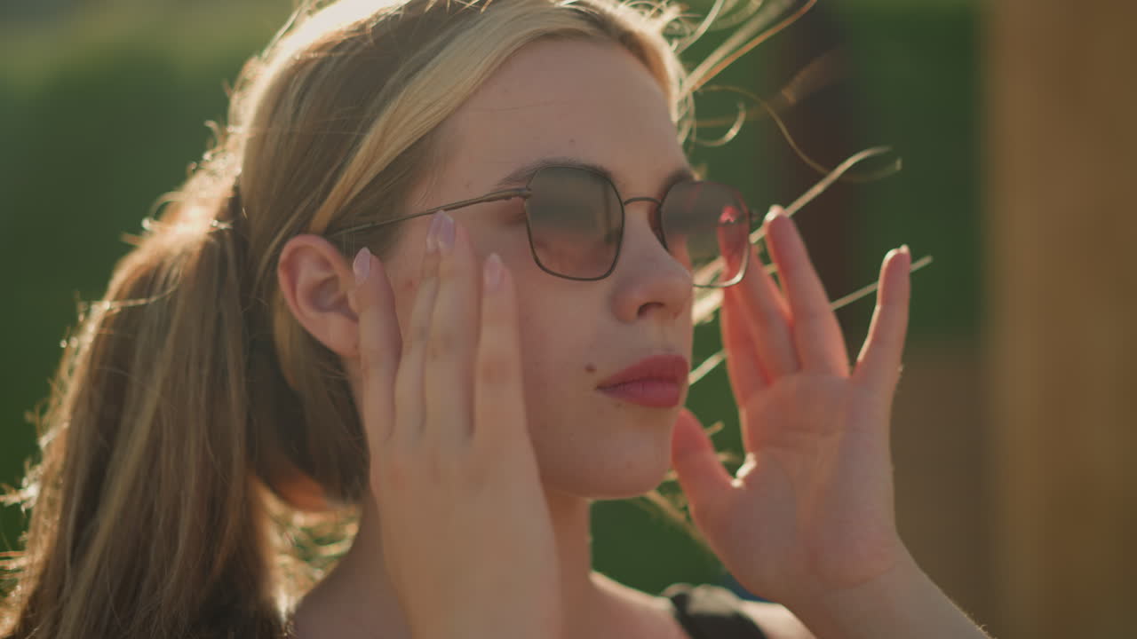 молодая леди настраивает волосы и солнцезащитные очки с серьезным выражением лица, пока ветер дует по ее волосам, солнечный свет отражается на ее лице, создавая спокойное свечение, размытый фон
