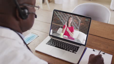 médico afroamericano que usa auriculares de teléfono tomando notas mientras hace una videollamada en una computadora portátil