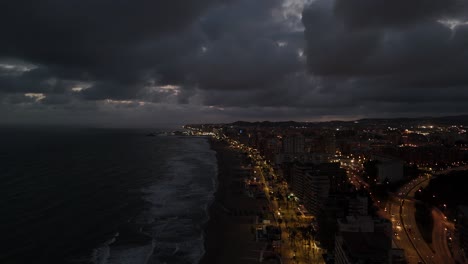 drone capturing the illuminated coastline of torreblanca, spain at dusk, with dark clouds creating a dramatic atmosphere over the mediterranean sea, pulling out shot