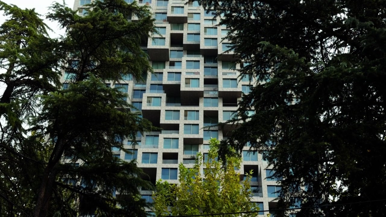 Una torre moderna con secciones cuadradas salientes se alza detrás de los árboles en Tirana, Albania. La vista cercana destaca la fachada geométrica y las filas de ventanas de cristal