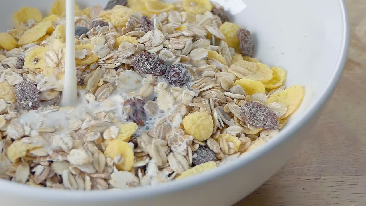 tiro deslizante en cámara lenta de verter leche en muesli en un tazón de cereal blanco en un mostrador de cocina para el desayuno