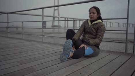 Serious-young-sportswoman-stretching-legs-on-jetty