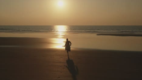 Eine-Silhouette-Einer-Glücklichen-Frau,-Die-Vor-Freude-Springt,-Während-Sie-Während-Eines-Wunderschönen-Goldenen-Sonnenuntergangs,-Indien,-An-Einem-Strand-Entlang-In-Richtung-Meer-Läuft