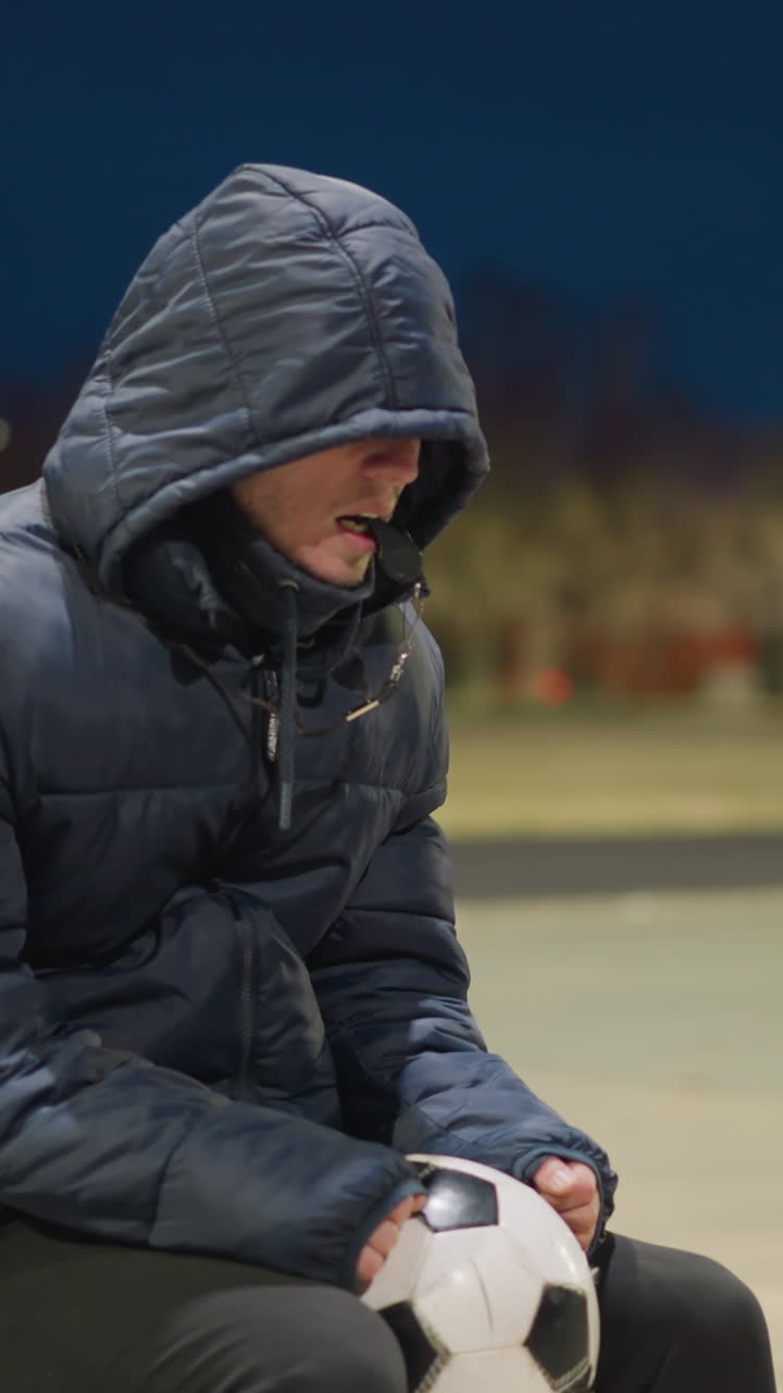 한 남자가 야외의 벤치에 앉아서 입에 휘파람을 치고 다리 사이에 축구공을 들고 있으며, 배경은 희미하게 밝고 흐릿하며, 멀리 떨어진 건물과 필드를 특징으로합니다.