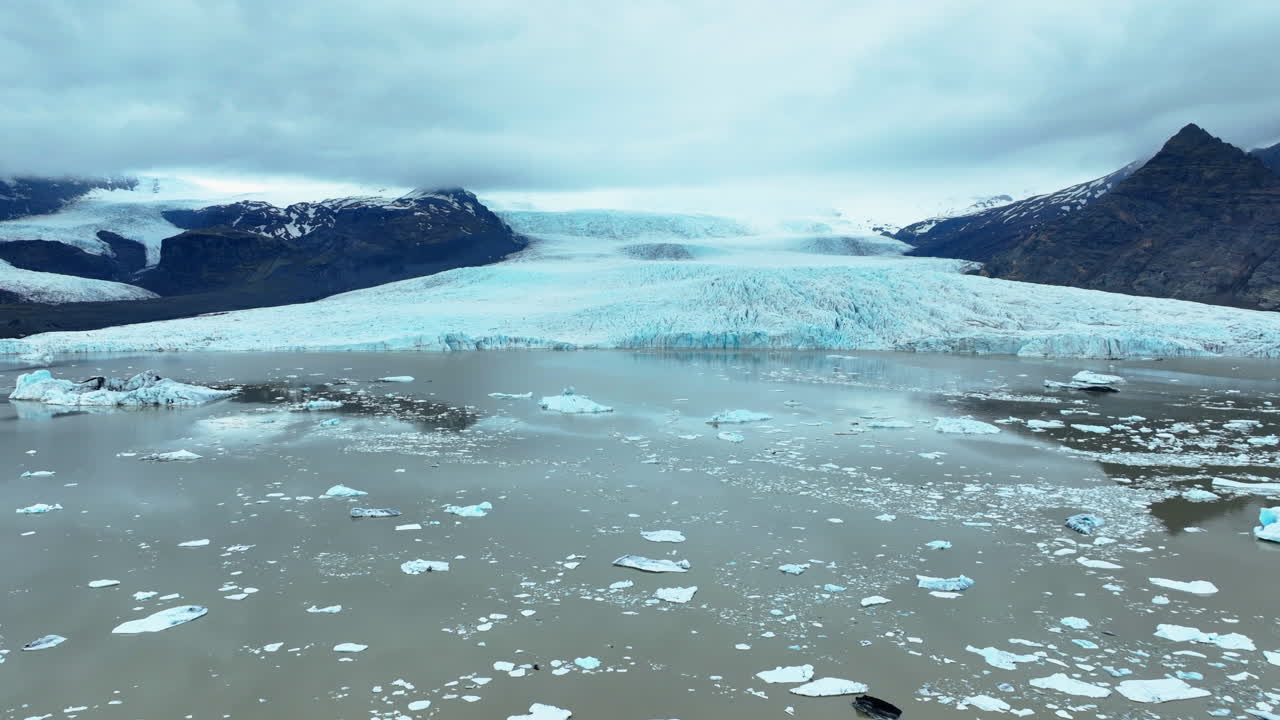 Massive icebergs drifting through blue gray glacial waters with Vatnajokull glacier looming in background, revealing stark environmental transformations