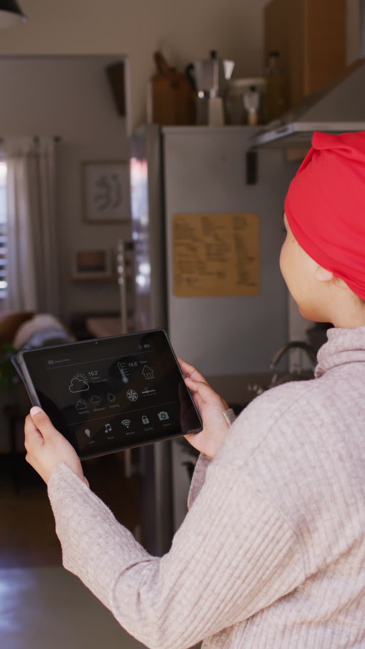 Vertical video of biracial woman in hijab at home holding tablet with smart home interface screen