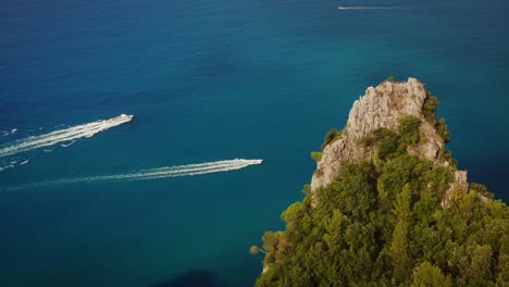 perfectly composed drone orbit shot of coastal rock formation with trees, small cruise ship and speedboat sailing vivid blue waters in the background