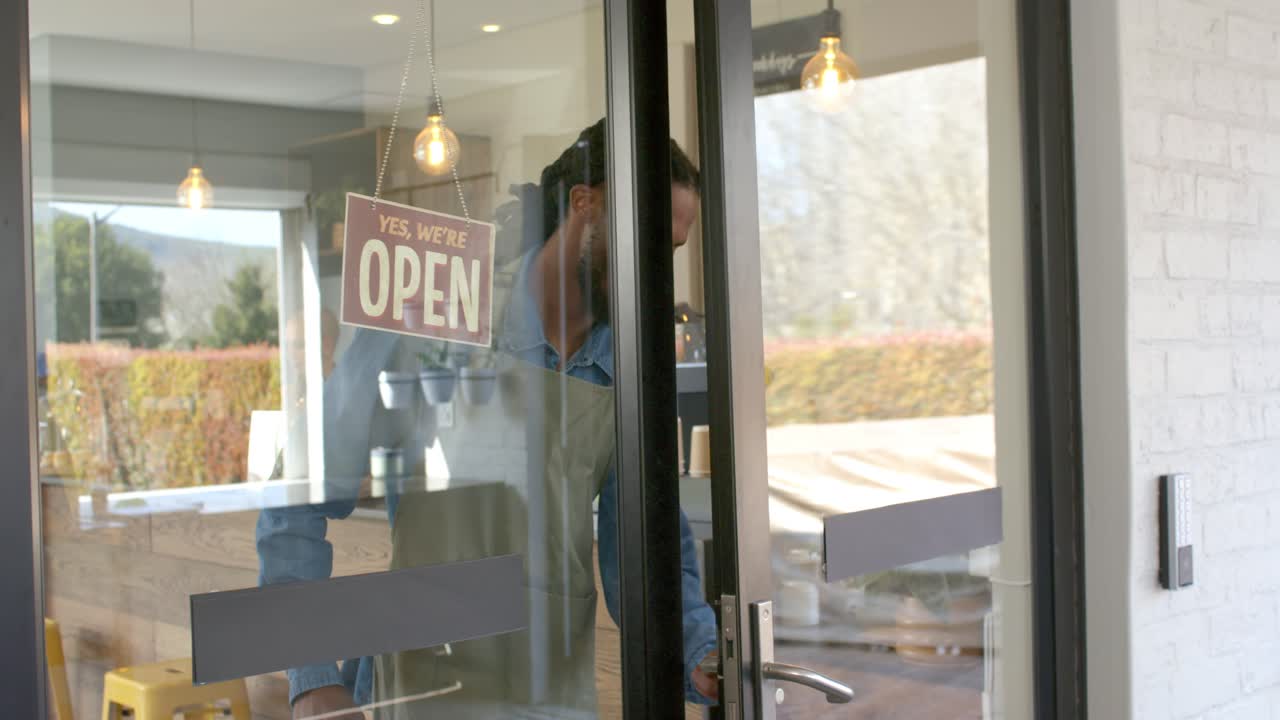 Homem afro-americano virando a placa de fechado para aberto em um café, abrindo a porta de vidro e dando as boas-vindas aos clientes